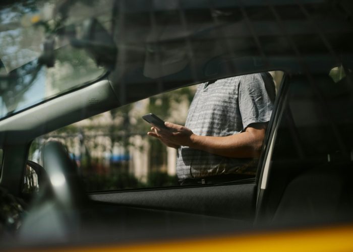A man using a smartphone inside a car, visible through the window in an urban setting.
